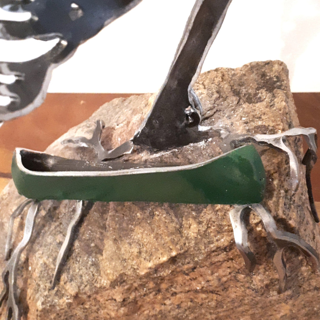 detail of metal green canoe on granite, under a metal tree sculpture