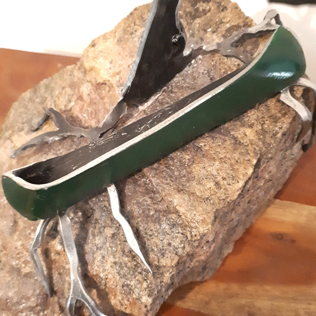 detail of metal green canoe on granite, under a metal tree sculpture