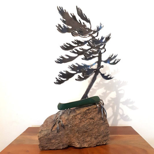 Metal sculpture of a green canoe under a metal white pine on a rock 